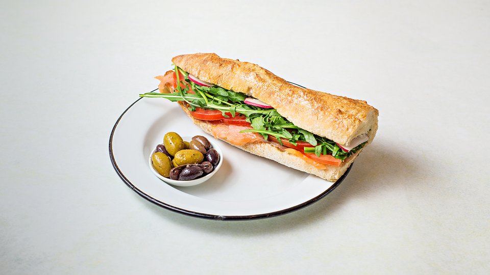 כריך בבגט בהיר עם עלים ירוקים ועגבנייה, לצד קערית זיתים